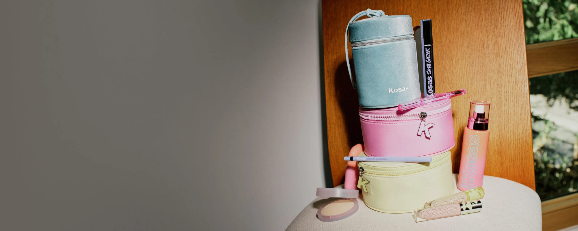 Kosas holiday cases stacked beside a spray bottle, a pressed powder and a concealer on a cream stool